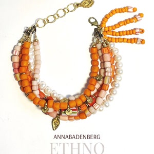 Può includere: Braccialetto multifilo con perline arancioni, rosa e bianche, un ciondolo dorato e una chiusura a catena dorata. Il braccialetto ha un fiocco di perline arancioni.