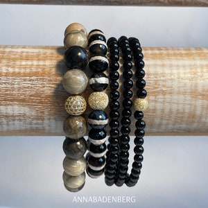 May include: A stack of bracelets featuring black onyx, brown and white agate, and gold beads. The bracelets are displayed on a wooden surface. The text "ANNABADENBERG ONYX" is visible at the bottom of the image.