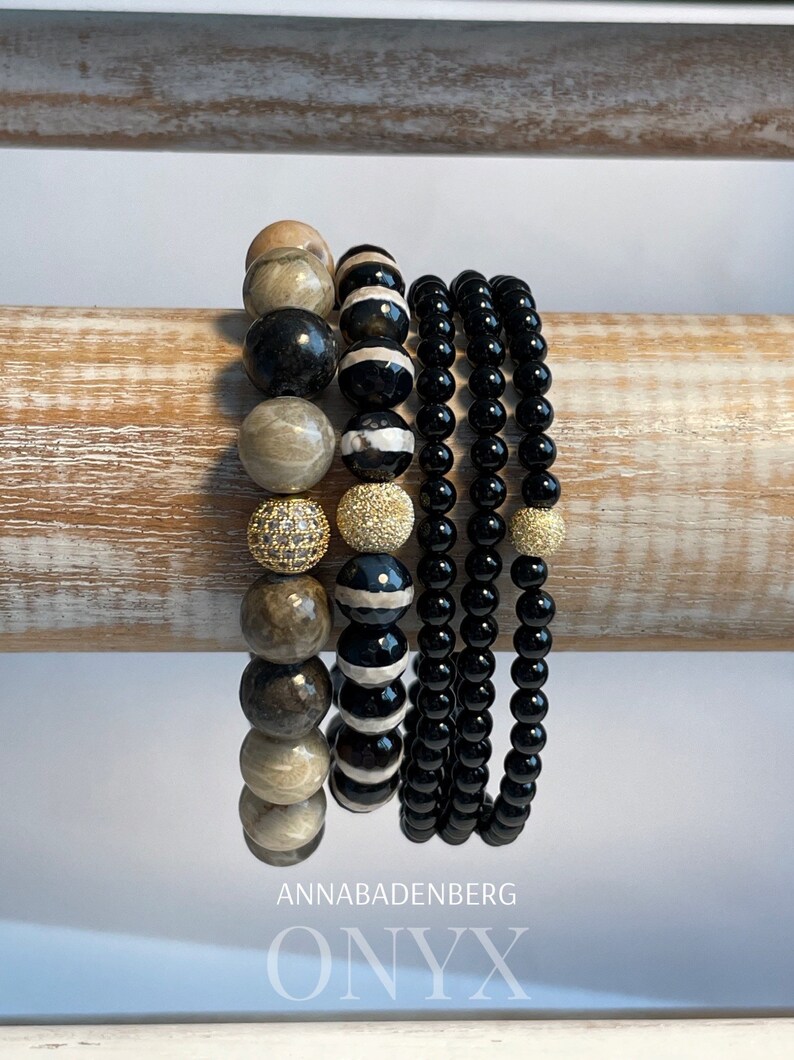 May include: A stack of bracelets featuring black onyx, brown and white agate, and gold beads. The bracelets are displayed on a wooden surface. The text "ANNABADENBERG ONYX" is visible at the bottom of the image.