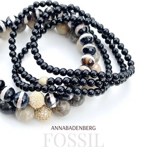 May include: A set of black and white agate gemstone bracelets with gold accents. The bracelets are stacked on top of each other and feature a variety of different patterns. The text "ANNABADENBERG FOSSIL" is visible at the bottom of the image.