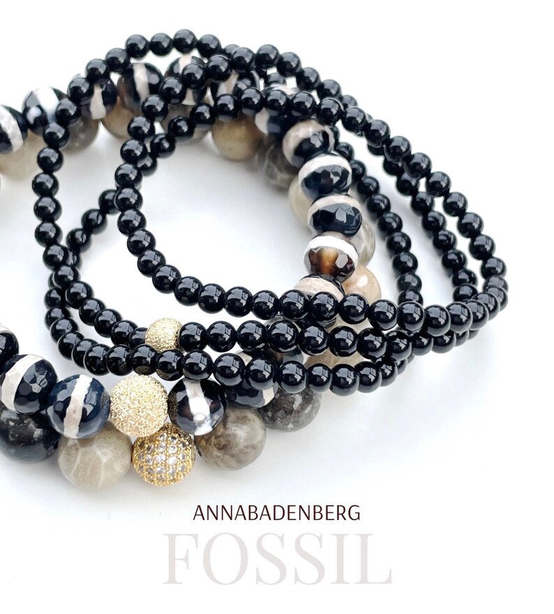 May include: A set of black and white agate gemstone bracelets with gold accents. The bracelets are stacked on top of each other and feature a variety of different patterns. The text "ANNABADENBERG FOSSIL" is visible at the bottom of the image.