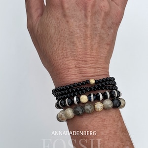 May include: A stack of bracelets with black, white, and gray beads. The bracelets feature gold accents and a small, clear crystal. The text "ANNABADENBERG" and "FOSSIL" is visible below the bracelets.