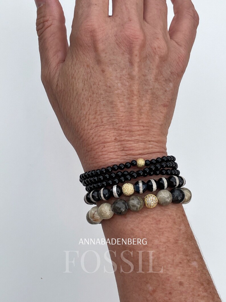 May include: A stack of bracelets with black, white, and gray beads. The bracelets feature gold accents and a small, clear crystal. The text "ANNABADENBERG" and "FOSSIL" is visible below the bracelets.