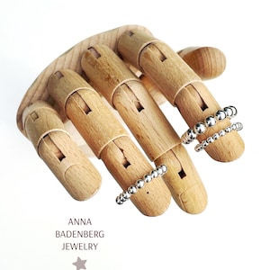 Può includere: Un manichino di mano in legno con tre braccialetti di perline argentate sulle dita. Il manichino di mano è su uno sfondo bianco. Il testo "ANNA BADENBERG JEWELRY" è visibile sotto il manichino di mano.