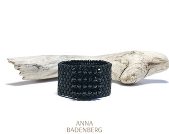 May include: A black beaded ring with a square design. The ring is on a piece of driftwood. The text "ANNA BADENBERG JEWELRY" and a star are below the ring.
