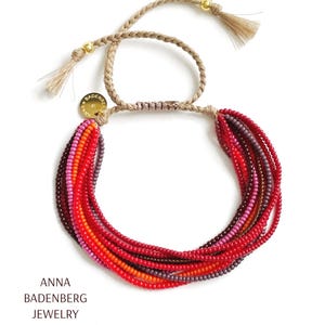以下が含まれることがあります： 赤、オレンジ、紫のビーズがグラデーションになった、マルチストランドビーズブレスレット。ブレスレットは編み紐と「Anna Badenberg Jewelry」と書かれたゴールドチャームでできています。