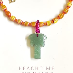May include: A beaded necklace with a green palm tree pendant. The necklace features orange and yellow beads, a pink accent, and a gold clasp. The text "BEACHTIME MADE BY ANNA BADENBERG" is at the bottom.