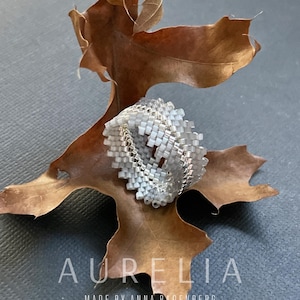 Op de afbeelding: Een zilveren en witte kralenring met een geometrisch patroon ligt op een bruin blad. De ring toont gedetailleerd kralenwerk. Het woord "AURELIA" is wit gedrukt, samen met de tekst "MADE BY ANNA BADENBERG".