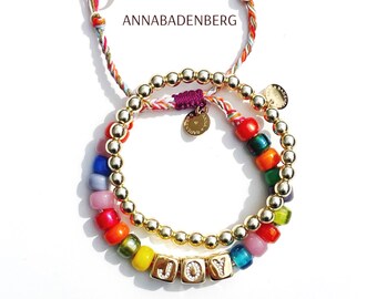 May include: Two gold beaded bracelets with colourful glass beads and a gold charm with the word "LOVE" on it. The bracelets are stacked on top of each other and spell out the word "JOY" with gold letter beads. The bracelets are tied with colourful strings with tassels.