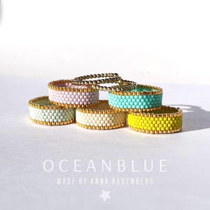 Può includere: Anelli di perline in vari colori pastello: rosa, verde menta, crema, turchese e giallo. Ogni anello ha un bordo dorato e sono disposti su una superficie bianca. Il testo "OCEANBLUE" e "MADE BY ANNA BADENBERG" sono visibili.