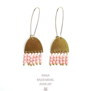May include: A pair of dangle earrings with a brass arch-shaped top and pink beaded fringe. The earrings have long, thin gold-colored ear wires. The text "ANNA BADENBERG JEWELRY" is printed below the earrings.