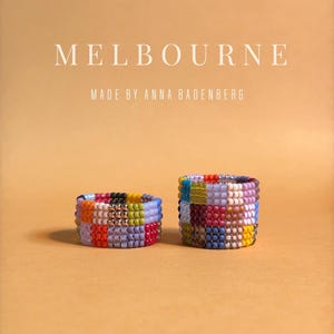 Könnte beinhalten: Zwei Ringe aus kleinen, bunten Glasperlen in einem Gittermuster. Über den Ringen steht der Text MELBOURNE und MADE BY ANNA BADENBERG. Die Perlen haben verschiedene Farben, darunter Rot, Blau, Gelb und Violett.