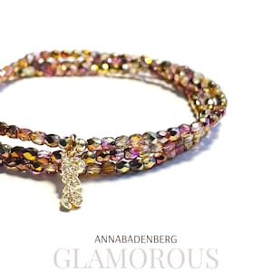 Könnte beinhalten: Ein mehrreihiges Armband mit goldenen und rosafarbenen, schillernden Perlen und einem kleinen goldenen Anhänger mit klaren Strasssteinen. Das Armband liegt auf einem weißen Hintergrund. ANNABADENBERG GLAMOROUS steht in schwarzer Schrift unter dem Armband.