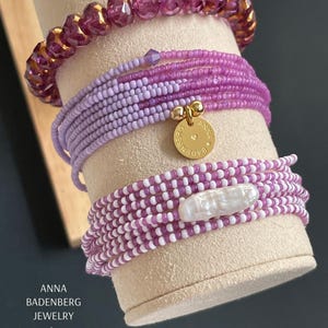 Könnte beinhalten: Ein Set aus lila und weißen Perlenarmbändern mit einem goldenen Anhänger, auf dem "Anna Badenberg Jewelry" steht.