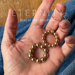 Peut inclure: Deux boucles d'oreilles créoles dorées composées de perles. Les boucles d'oreilles sont circulaires, chacune étant composée de petites sphères espacées uniformément. Le texte "ETERNITY MADE BY ANNA BADENBERG" est visible.