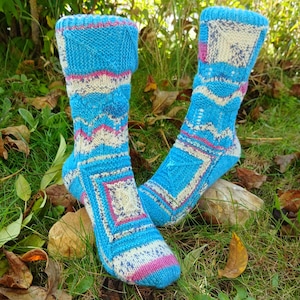 Puede incluir: Un par de calcetines tejidos a mano en tonos turquesa, blanco y rosa. Los calcetines presentan patrones geométricos y se exhiben sobre hierba y hojas. Un accesorio cálido.