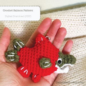 May include: A handmade crochet salmon keychain in red, green, and white. The fish has a detailed design with white accents and a small metal ring for attachment. The text "Crochet Salmon Pattern" and "Digital Download (PDF)" is in the upper left corner.