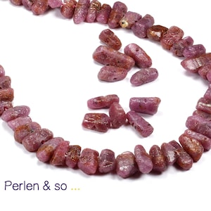 Könnte beinhalten: Eine Halskette aus rohen, rötlich-rosa Rubinkugeln. Die Perlen sind unregelmäßig geformt und aufgefädelt, mit einigen losen Perlen, die herumliegen. Der Text "Perlen & so ..." befindet sich unten.