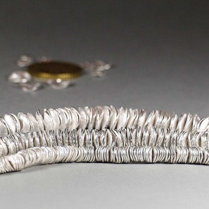 May include: Close-up of two strands of silver-colored disc-shaped beads. The beads have a textured surface and are strung together. In the background, a blurred silver necklace with a gold-colored pendant is visible.
