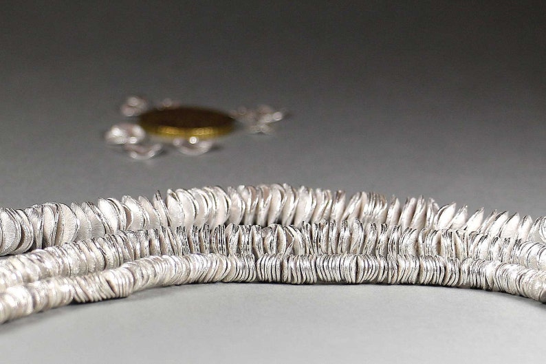 May include: Close-up of two strands of silver-colored disc-shaped beads. The beads have a textured surface and are strung together. In the background, a blurred silver necklace with a gold-colored pendant is visible.