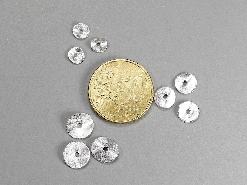 May include: A collection of small, silver-colored, disc-shaped beads with a textured surface and a central hole. A gold coin with the number 50 is next to the beads. The beads are scattered on a gray background.
