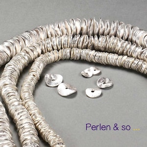 Könnte beinhalten: Silberfarbene, scheibenförmige Perlen, die zu mehreren Strängen aufgereiht sind. Mehrere lose Perlen sind in der Nähe verstreut. Die Perlen haben eine strukturierte Oberfläche. Der Text "Perlen & so ..." ist sichtbar.