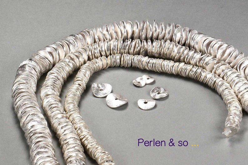 May include: Silver-colored, disc-shaped beads strung into multiple strands. Several loose beads are scattered nearby. The beads have a textured surface. The text "Perlen & so ..." is visible in the image.