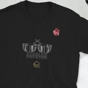 以下が含まれることがあります： 黒いTシャツに、白い鋼鉄製のドラム2つのグラフィックと、その下に「Steeles' Pots & Pans」というテキストが書かれています。