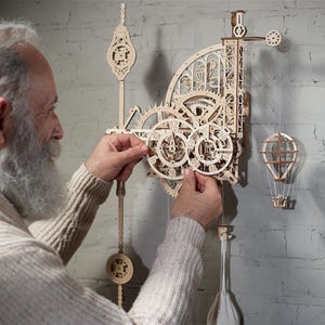 Op de afbeelding: Een persoon assembleert een houten klokset met ingewikkelde tandwielen en een ontwerp met een luchtballon. De klok is gemaakt van natuurlijk hout en heeft een rustieke uitstraling.