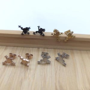 Könnte beinhalten: Eine Sammlung von Frosch-Ohrsteckern in verschiedenen Metallausführungen, darunter Schwarz, Gold, Roségold und Silber. Die Ohrringe werden auf einer Holzoberfläche präsentiert, die ihre verschiedenen Farben und Designs zeigt. Diese Ohrringe sind ein lustiges Accessoire.