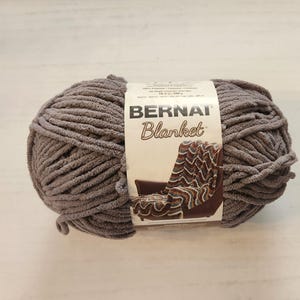 以下が含まれることがあります： グレーのベルナットブランケットヤーンの糸巻き。糸は柔らかくふわふわで、毛布、マフラー、その他の居心地の良いアイテムを作るのに最適です。糸巻きのラベルには「ベルナットブランケット」と書いてあります。