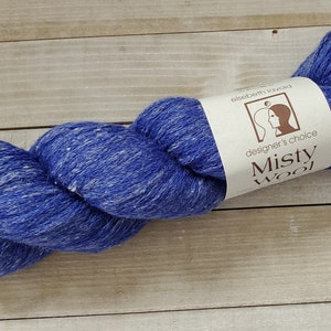 May include: A ball of blue and white speckled Misty Wool yarn. The yarn is tightly wound and has a label with the text "designer's choice Misty Wool". The yarn is on a light wood surface.