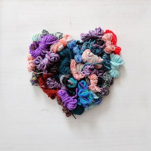 Puede incluir: Un arreglo en forma de corazón de hilos de colores en tonos morado, azul, rojo y turquesa. Los hilos tienen diferentes texturas y grosores, creando una exhibición visualmente atractiva. El fondo es una superficie blanca lisa.