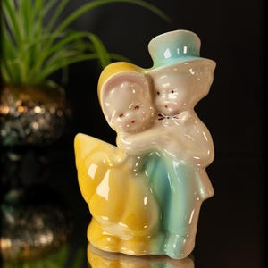 May include: A vintage ceramic figurine of a couple embracing. The man wears a light blue suit and top hat, while the woman is in a yellow dress and bonnet. The figurine has a glossy finish and is reflected on a dark surface.