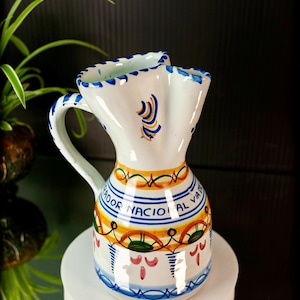 May include: White ceramic pitcher with a handle, decorated with blue, yellow, and orange bands and patterns. The pitcher has a flared top with a scalloped edge. Text on the pitcher reads "ADOR NACIONAL AL VIA".