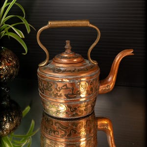 May include: Copper teapot with brass handle and spout. The teapot features decorative etched patterns in gold and copper tones. The teapot is sitting on a reflective surface, with a plant and decorative sphere in the background.