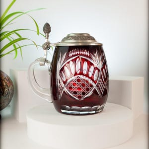 May include: A ruby red, cut-glass beer stein with a silver-tone hinged lid and handle. The glass features intricate geometric patterns and a clear glass handle. The stein is displayed on a white pedestal, showcasing its elegant design.