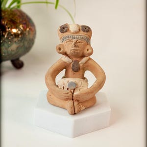 May include: A small, tan-colored figurine of a seated figure with a detailed headdress and ear ornaments. The figure is seated on a white block, with arms crossed and hands holding an object. The figurine is likely a representation of an ancient deity or important person.