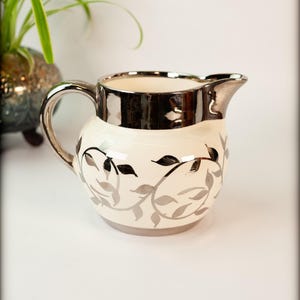 May include: Cream-colored ceramic pitcher with a silver rim and handle. The pitcher features a gray and black leaf design. The spout is silver. A decorative piece for serving beverages.