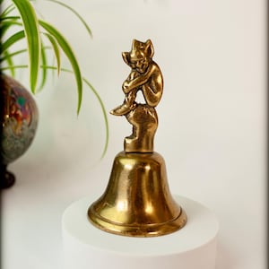 May include: A vintage brass bell with a seated figurine on top. The bell has a flared base and a small figure of a seated creature. The bell is displayed on a white pedestal.