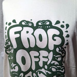 May include: White t-shirt with a green graphic of the words "FROG OFF!" surrounded by cartoon frogs.
