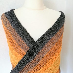 Puede incluir: Un chal de ganchillo con un diseño de cuello en V en tonos naranja, marrón y negro. El chal está drapeado sobre un maniquí blanco.