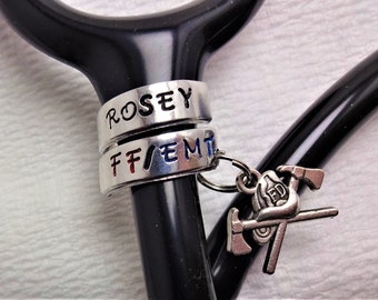May include: A black stethoscope with two silver rings engraved with the names "Rosey" and "FF/EMT". A silver charm with a firefighter helmet, crossed axes, and the letters "FD" hangs from the stethoscope.
