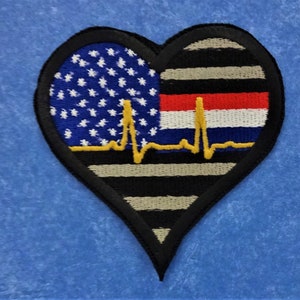 Puede incluir: Un parche en forma de corazón con una bandera estadounidense en el lado izquierdo y una bandera de línea azul delgada en el lado derecho. Una línea de ECG amarilla está centrada en el parche.