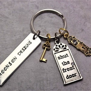 Op de afbeelding: Een zilveren sleutelhanger met een zilveren tag waarop "MOONSON DESIGNS" staat en een zilveren hanger met de tekst "sluit de voordeur". De sleutelhanger heeft ook een gouden sleutelhanger en een gouden deurknophanger.