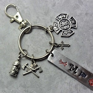 May include: Silver keychain with a variety of charms, including a fire hydrant, crossed axes, a firefighter's helmet, a cross, a firefighter's emblem, and a personalized tag with the letter "M" and a fire department logo.