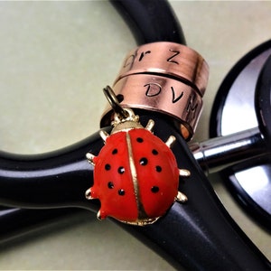 May include: A red ladybug charm with black spots and gold accents attached to a stethoscope with three copper rings engraved with "Dr Z" and "DVM".