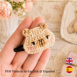 Puede incluir: Pinza de pelo de oso de ganchillo en color beige, con ojos y nariz negros y mejillas rosas. La pinza está sujeta a una horquilla negra. El texto "PDF Pattern in English & Español" es visible.