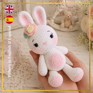 Puede incluir: Un conejito blanco tejido a crochet con detalles rosas en las orejas, mejillas y vientre. El conejito tiene ojos negros y una flor en la cabeza. La imagen incluye el texto "Pattern in English & Español".