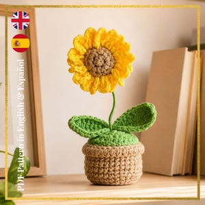 May include: A crocheted sunflower in a pot. The sunflower has yellow petals and a brown center. The green stem supports the flower and two green leaves. The pot is tan and sits on a wooden surface. The text "PDF Pattern in English & Español" is visible.